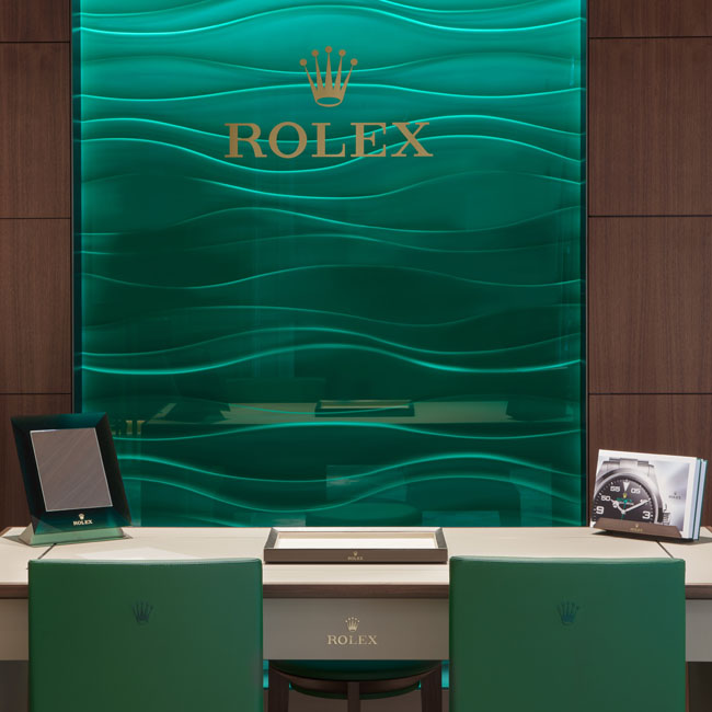 Grünes Glaselement mit goldenem Rolex Logo an dunkler Wand. Davor ein Tisch mit zwei Stühlen.